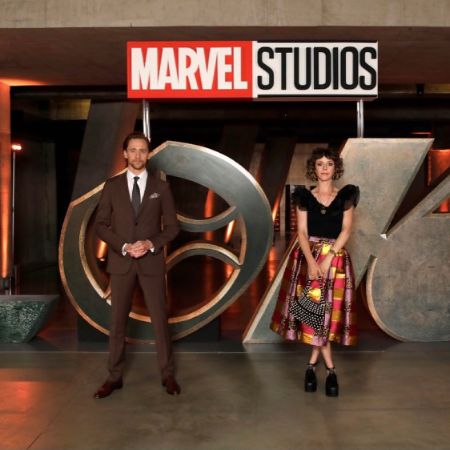 Sophia Di Martino and Tom Hiddleston are standing next to each other with Loki logo in the background.